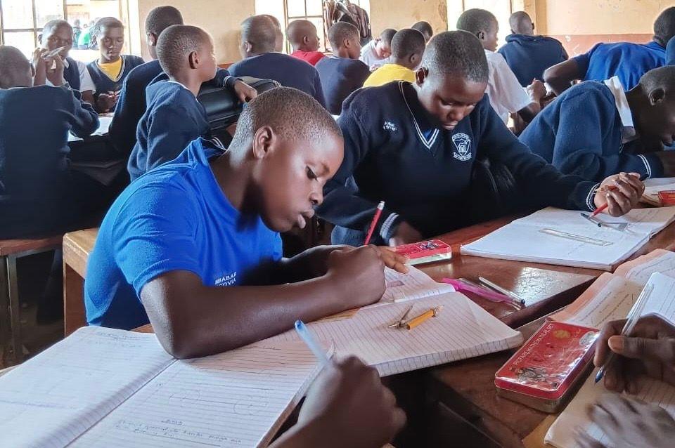 Students in a classroom taking notes, sitting in groups at shared desks with notebooks and pencils.