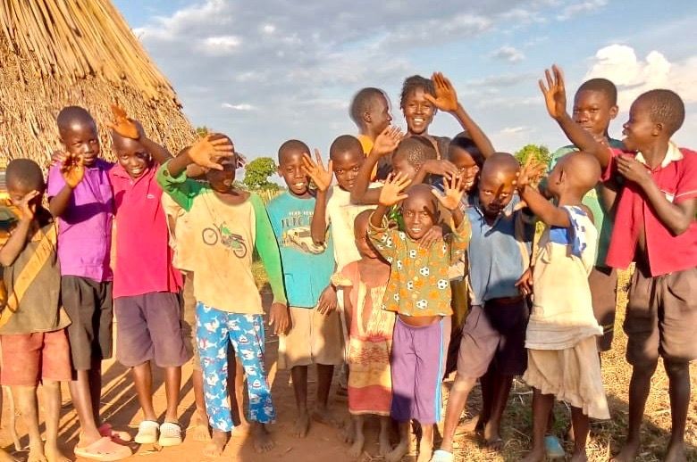 Group of children smiling and waving in front of a thatched hut and cloudy sky background.