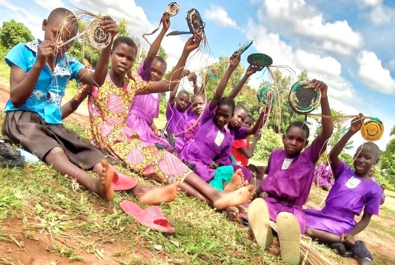 Group of children sitting outdoors holding up woven crafts, smiling under a partly cloudy sky.