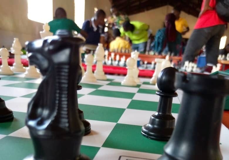 Close-up of a chessboard with black pieces in focus, while people gather in the background.
